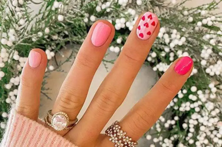 Hand showcasing a variety of pink Valentine’s Day nail designs, adorned with elegant rings against a backdrop of white flowers.